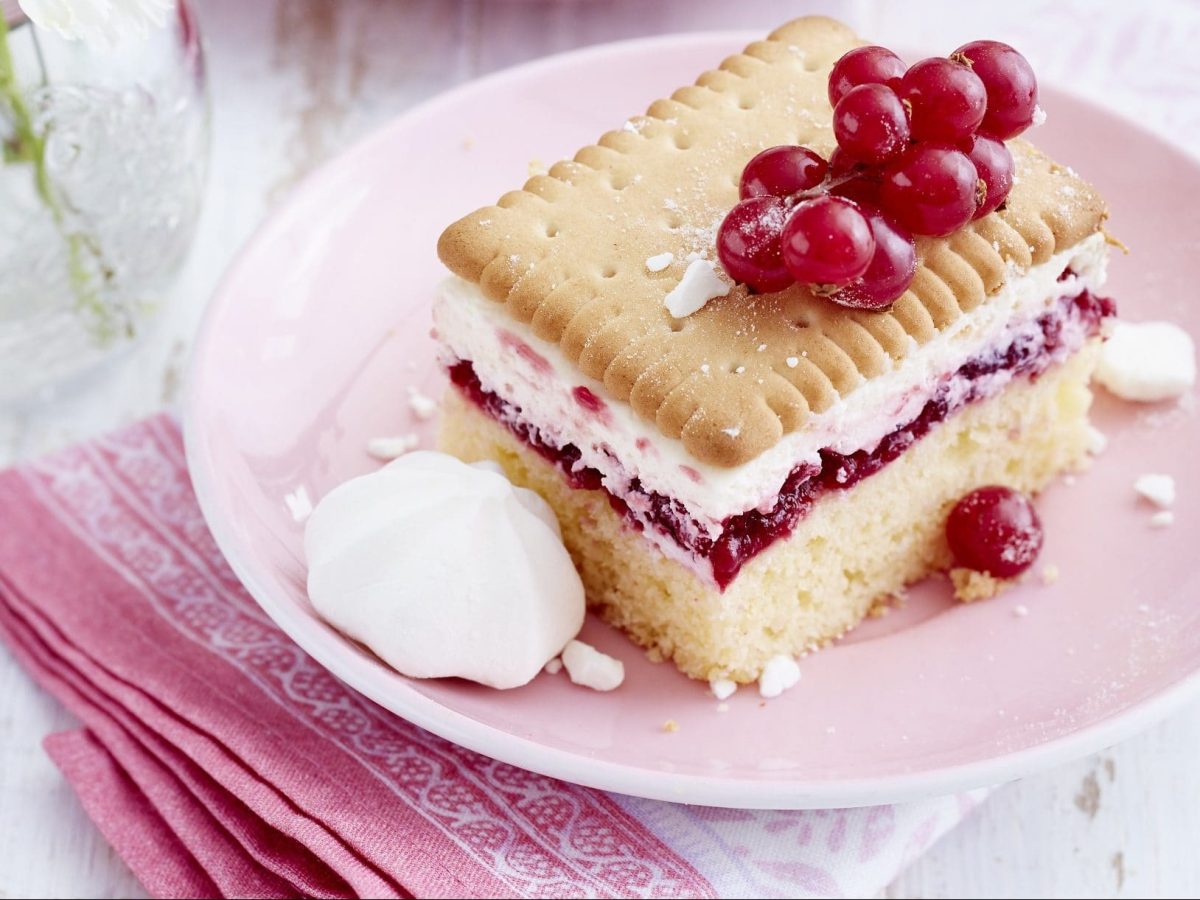 Ein Stück Butterkekskuchen mit Johannisbeeren angerichtet auf einem rosa Teller und mit rosa Serviette und Blumen als Deko im Hintergrund.