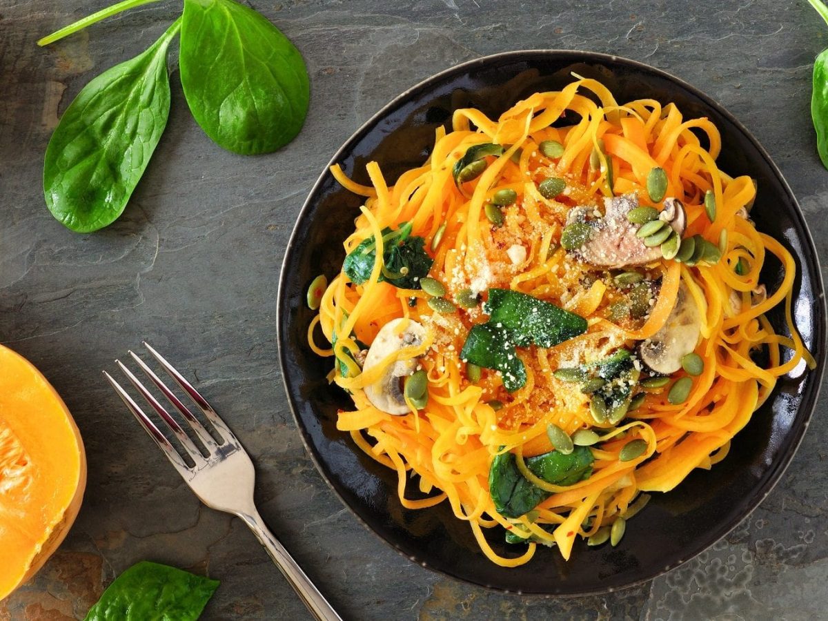 Eine Portion Kürbispasta mit Champignons, Kürbiskernen und Parmesan steht auf einem schwarzen Teller auf einem Untergrund aus Stein. Am linkenRand liegt ein Butternut-Kürbis. Um den Teller herum liegen einzelne Spinatblätter.