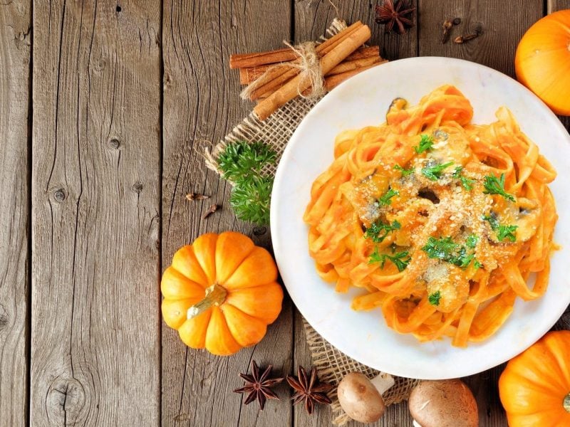 Unsere Butternut-Pasta mit Champignons auf einem weißen Teller. Die Pasta ist angerichtet mit Parmesan und Petersilie. Der Teller steht auf einem braunen Untergrund.