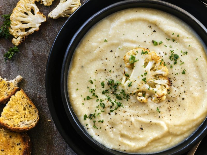 Die indische Blumenkohlsuppe füllt die schwarze Schüssel randvoll aus. Dazu gibt es etwas gerösteten Blumenkohlröschen als Garnitur und Röstbrot.