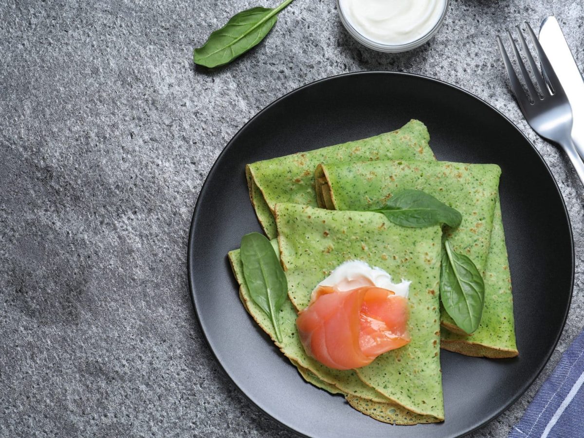 Drei Spinat-Pfannkuchen liegen zusammengeklappt auf einem schwarzen Teller. Dazu Crème fraîche und Lachs als Topping.