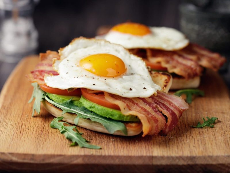 Die Avocado-Ei-Bacon-Stulle rustikal angerichtet auf einem Holzbrett.