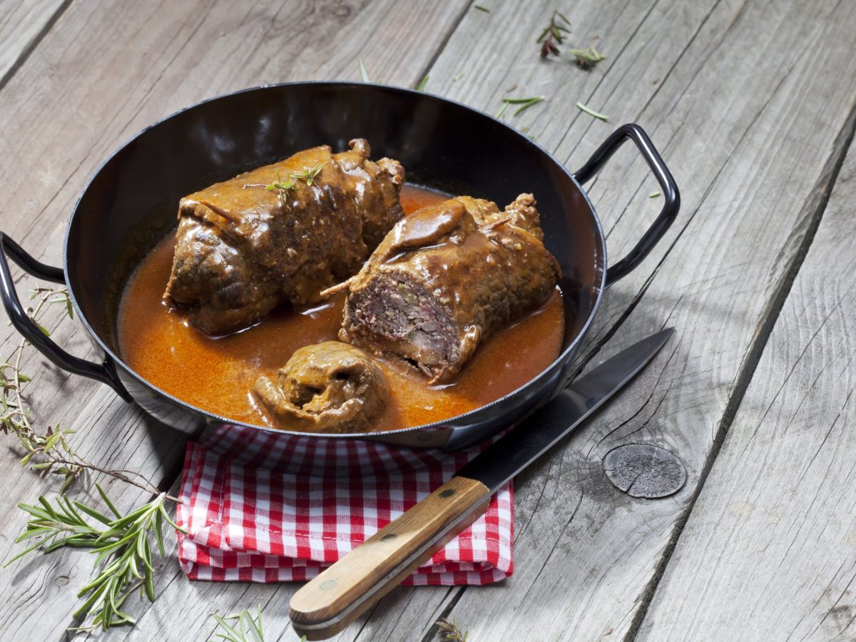 Rinderrouladen in gusseiserner Pfanne mit Soße auf grauem, hölzernem Untergrund.