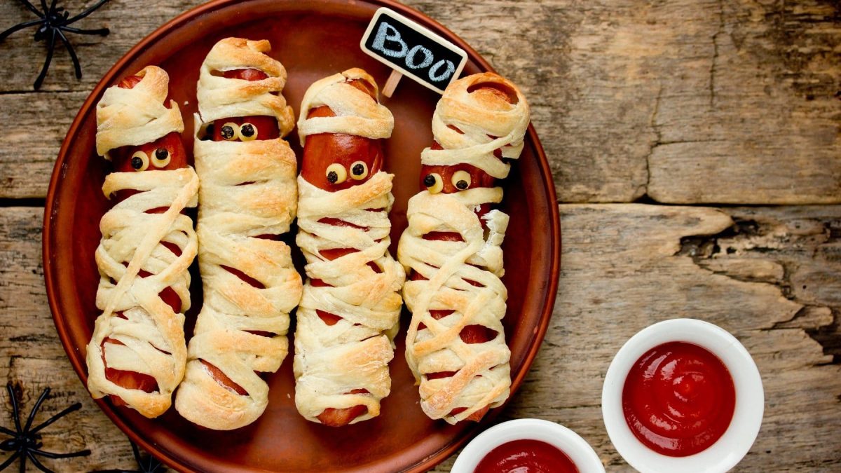 Mumien-Würstchen auf einem braunen Teller. Die Würstchen haben Pfefferkörner als Augen und am Rand des Tellers stehen zwei Schalen mit Ketchup.