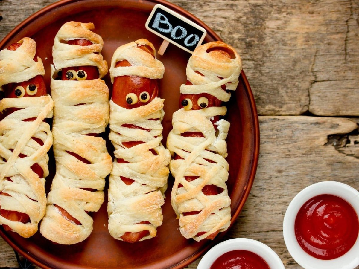 Mumien-Würstchen auf einem braunen Teller. Die Würstchen haben Pfefferkörner als Augen und am Rand des Tellers stehen zwei Schalen mit Ketchup.