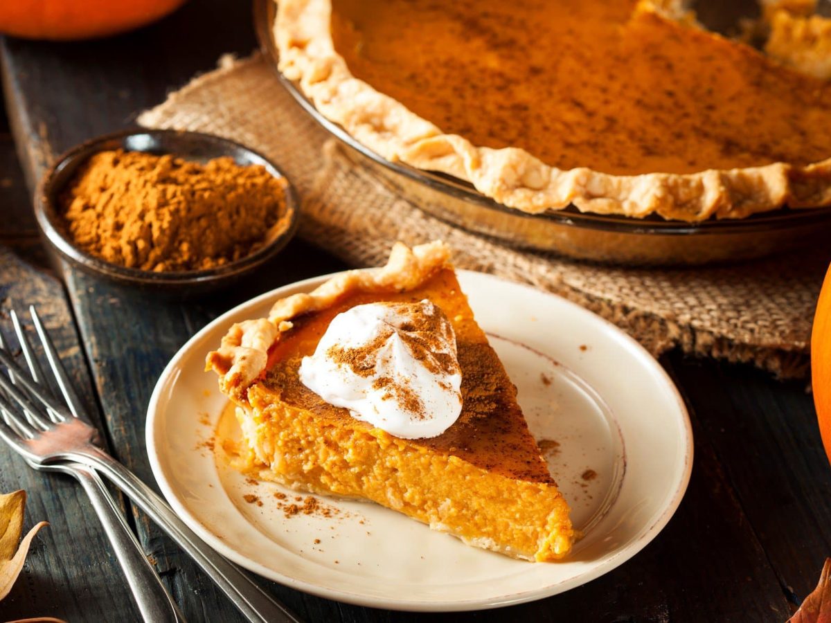 Angeschnittener Kürbiskuchen auf weißem Teller. Umgeben vom ganzen Kuchen und herbstlicher Dekoration.