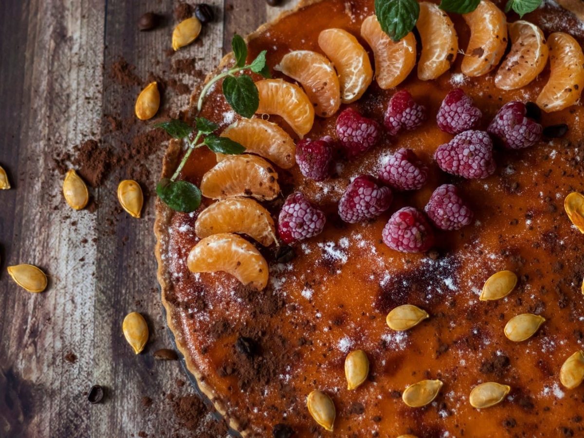 Ein Pumpkin Pie mit Obst ist angerichtet mit Mandarinen, Himbeeren, Kakaopulber und Puderzucker. Auf dem Kuchen und dem Untergrund liegen Kürbiskerne verteilt. Der Untergrund beseht aus braunem Holz.