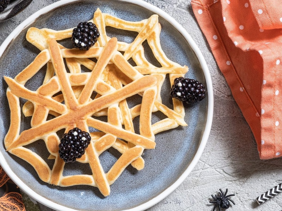 Zwei Spinnennetz-Pfannkuchen mit Brombeeren auf einem gemusterten Teller auf einem grauen Tisch. Um den Teller herum liegen Servietten, ein geschirrtuch, eine Schale mit Brombeeren und eine Plastik-Spinne. Außerdem liegen am rechten Bildrand zwei gemusterte Strohhalme.