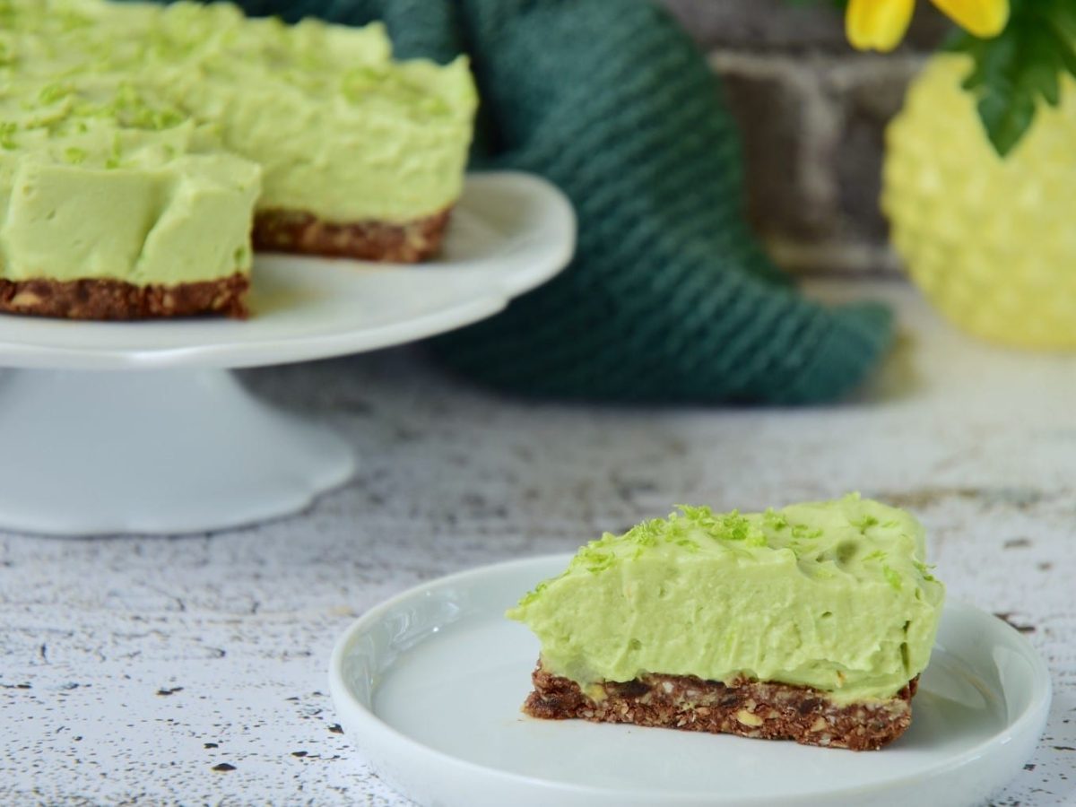 Ein Stück Avocado-Cheesecake mit Pistazien steht auf einem hellgrünen Teller auf einem gemusterten Untergrund. Im Hintergrund steht der restliche Kuchen auf einem Kuchentablett.