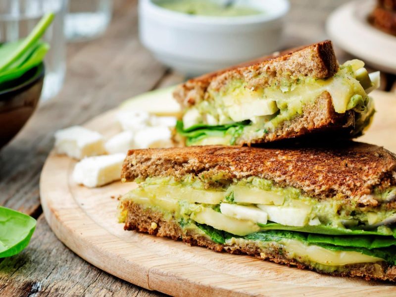 Auf einem Holzbrett liegen Avocado-Toasties, drumherum sieht man unscharf Zutaten wie Babyspinat, Käsewürfel oder Pesto.