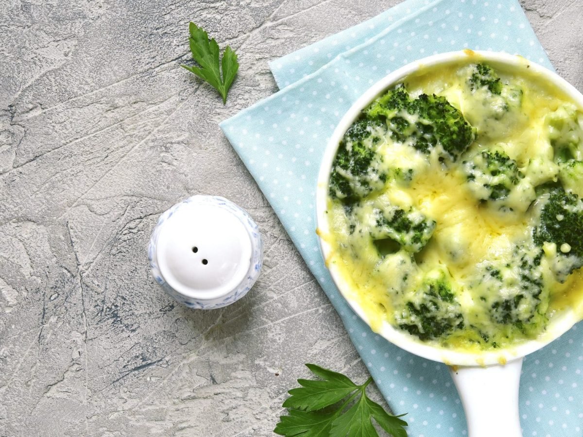 Das geschichtete Brokkoli-Blumenkohl-Gratin in einer Auflaufform. Dazu steht auf dem Tisch ein Salzstreuer, eine Gabel und ein Glas Wasser mit Zitrone.