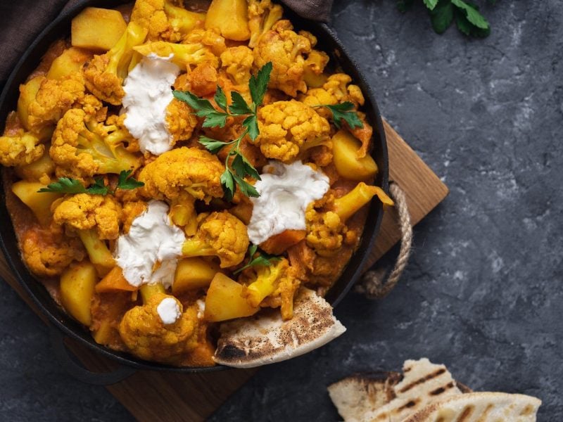 EIn Topf mit Blumenkohl-Curry und Joghurt, daneben liegt etwas Naanbrot.