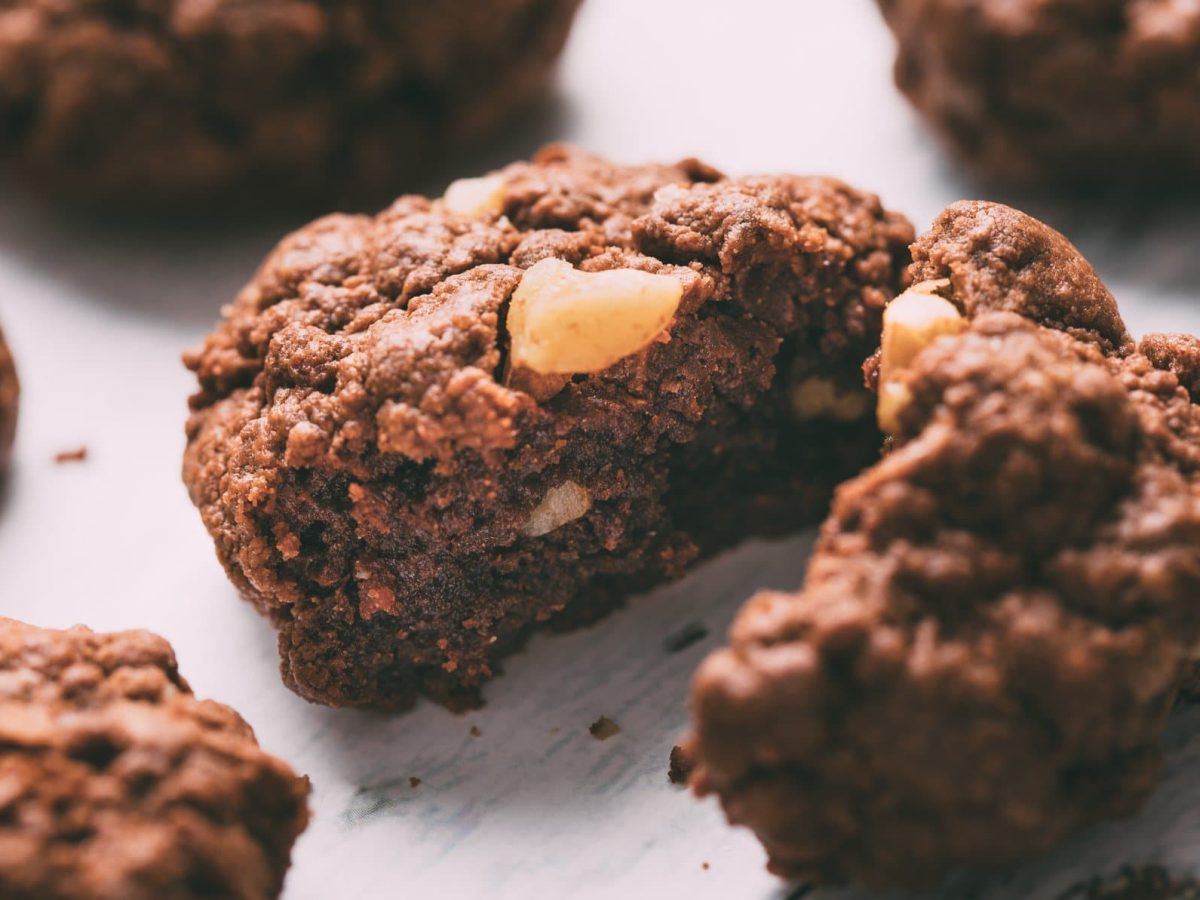 Ein angeschnittener Chocolate Chip Cookie mit dreierlei Schokolade auf hellem Holzuntergrund, drumherum sieht man unscharf ein paar weitere Kekse.