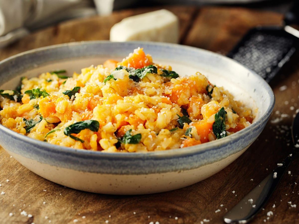 Ein tiefer Teller mit Erdnuss-Möhrenrisotto, verziert mit frischen Kräutern und Parmesan, auf einem Holztisch. Daneben liegt ein dunkler Löffel, im Hintergrund ist unscharf ein Stück Käse und eine Reibe zu sehen.