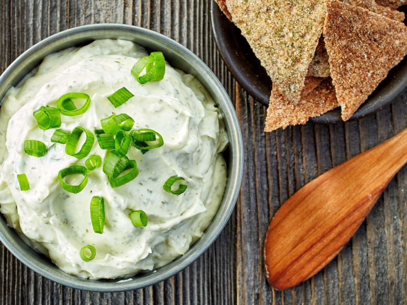 In der Draufsicht: eine Schale Feta-Minz-Dip mit Frühlingszwiebel bestreut, daneben sieht man ein Holz-Buttermesser und ein paar Cracker.