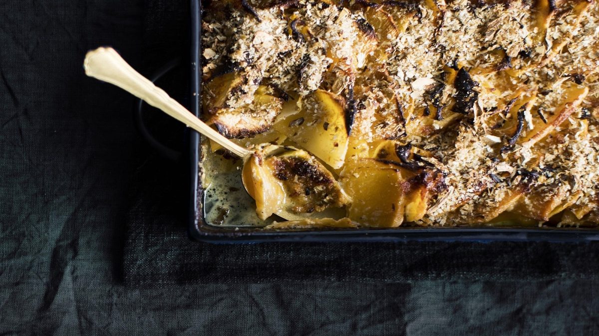 Die Ecke einer Auflaufform gefüllt mit gebackenem Kohlrabi mit Apfelringen mit KÄse überbacken. Ein Löffel liegt in dem angebrochenen Gericht. Vor einem dunklem Hintergrund, von oben.