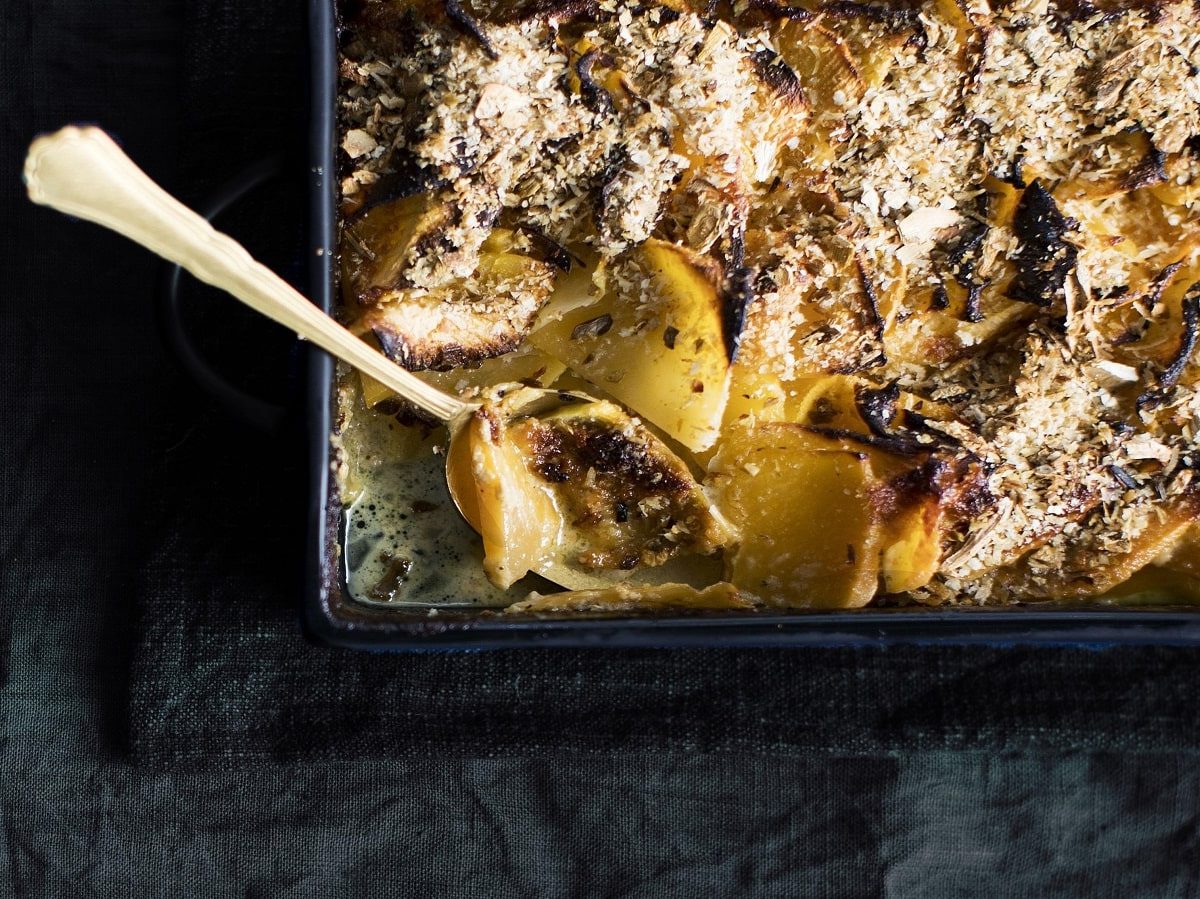 Die Ecke einer Auflaufform gefüllt mit gebackenem Kohlrabi mit Apfelringen mit KÄse überbacken. Ein Löffel liegt in dem angebrochenen Gericht. Vor einem dunklem Hintergrund, von oben.