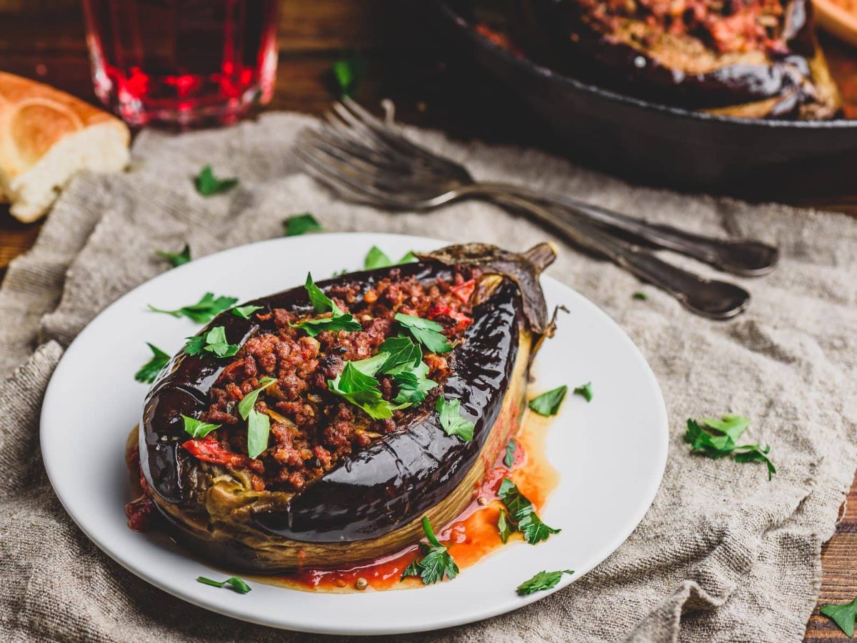 Auf einem weißen Teller liegt eine gefüllte Aubergine mit Nussmus, darüber gestreut ist Petersilie. Drumherum liegt etwas Brot, Besteck sowie eine Pfanne mit weiteren Auberginen.