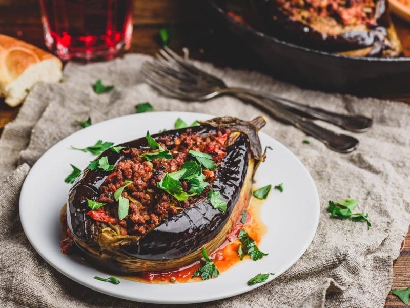 Auf einem weißen Teller liegt eine gefüllte Aubergine mit Nussmus, darüber gestreut ist Petersilie. Drumherum liegt etwas Brot, Besteck sowie eine Pfanne mit weiteren Auberginen.