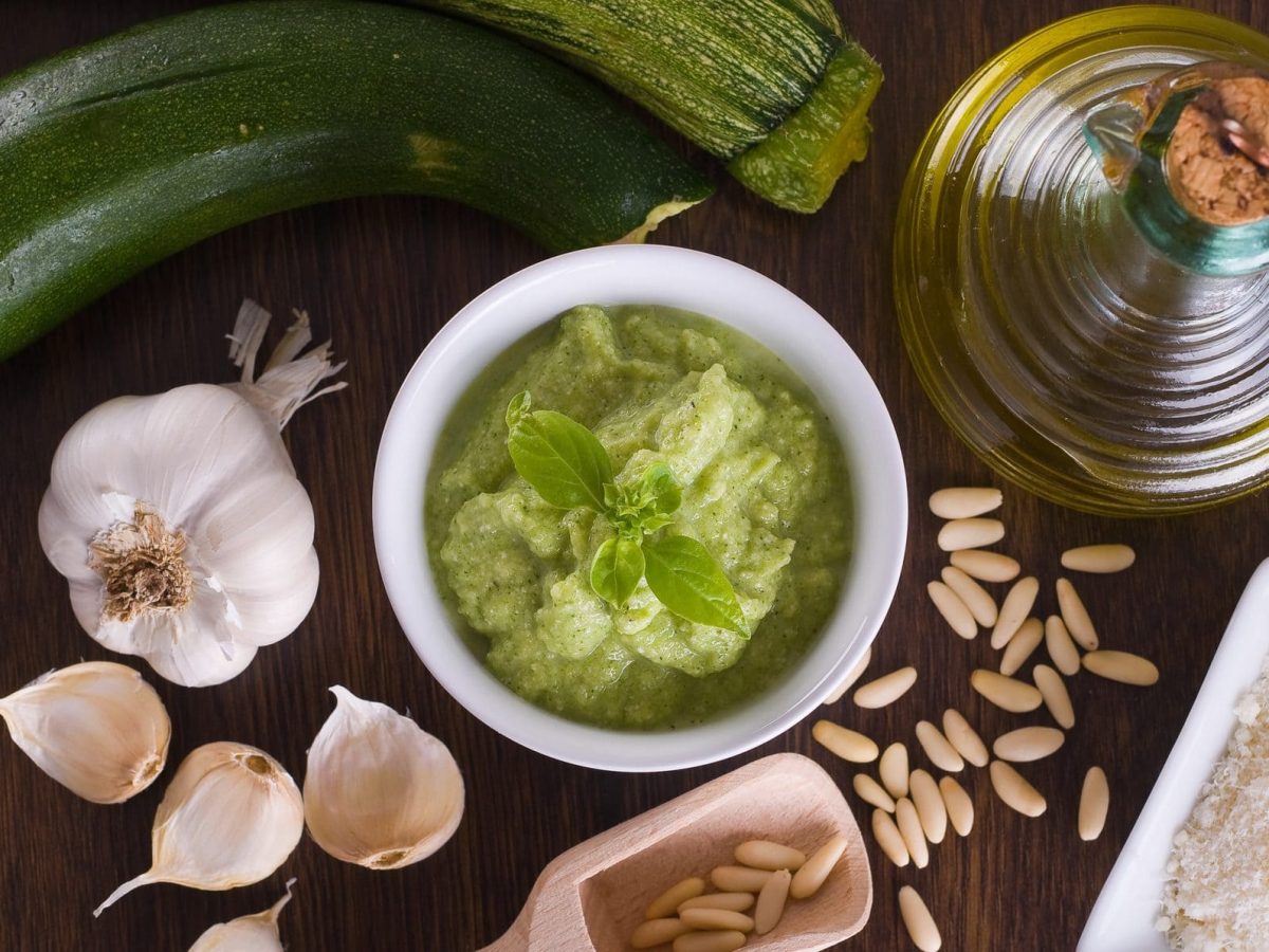 Auf einer Holzplatte sind zu sehen: eine Knoblauchknolle, eine Karaffe mit Olivenöl, zwei Zucchini, Pinienkerne, Salt, sowie eine runde Schale Zucchini-Pesto.