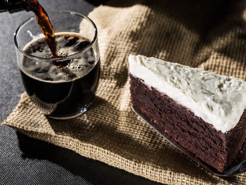 Ein Stück Guinness-Cake auf einem Kuchenheber neben einem Glas, in das gerade Guinness eingegossen wird, beides auf einem groben, braunen Tuch vor einem dunklen Hintergrund.