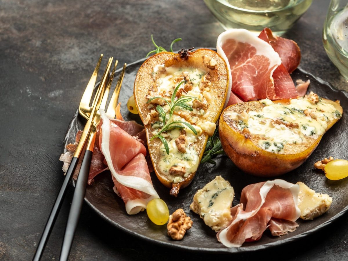 Auf einem rustikalen Tisch steht ein Teller, auf dem halbierte Birnen liegen. Diese sind mit Käse gefüllt. Neben den Birnenhälften liegen mehrere Scheiben Speck. Auf dem Teller liegen auch noch Walnüsse. Das Gericht heißt Birne mit Gorgonzola und Speck