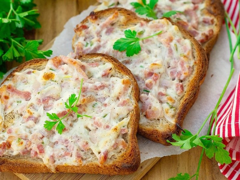 Drei Scheiben Toast sind mit einer Zwiebel-Speck-Mischung bestrichen. Sie liegen auf einem Papier und dann auf einem Holzbrettchen. Links ist Petersilie und rechts ein rot-weiß-gestreiftes Handtuch.