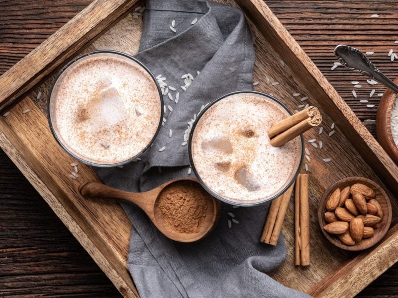 In der Draufsicht: ein Holztablett, auf dem zwei Gläser Horchata mit Eis und Zimtstangen stehen. Drumherum sind Zutaten wir Zimt, Mandeln und Reis verteilt.