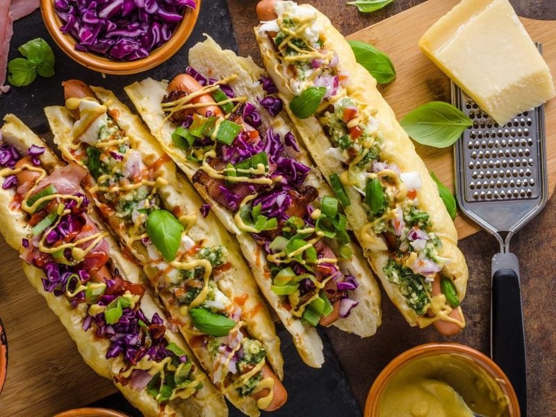 Hot Dogs mit Gemüse und Curry-Sauce auf einem Holzbrett. Die Hot Dog Brötchen sind mit Käse überbacken. Um die Hot Dogs herum liegt Bacon, vier Schalen mit Gurken, Lauch, Rotkohl und Curry-Sauce. Rechts neben den Brötchen liegt ein Stück Käse mit einem Käsehobel. Über den Hot Dogs ist Basilikum verteilt.