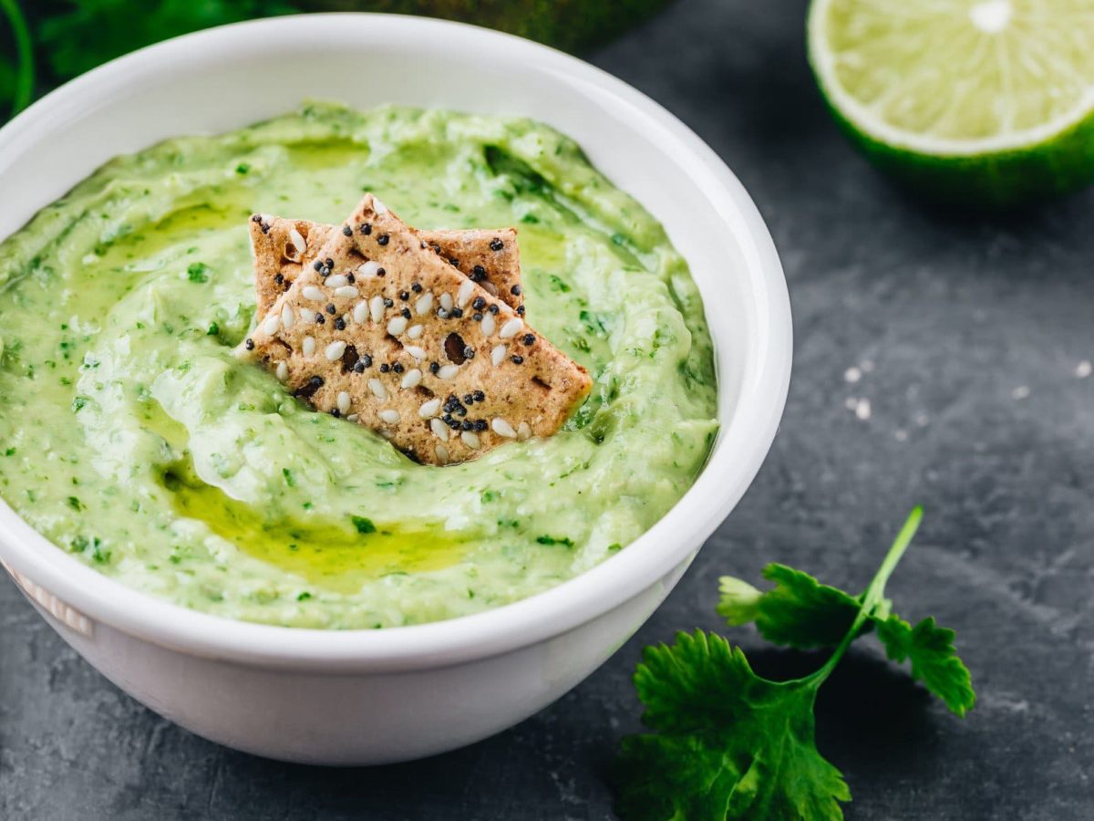 Eine weiße Schale mit Hummus mit Koriander, darin stecken ein paar Cracker. Daneben sieht man frischen Koriander sowie eine aufgeschnittene Limette.