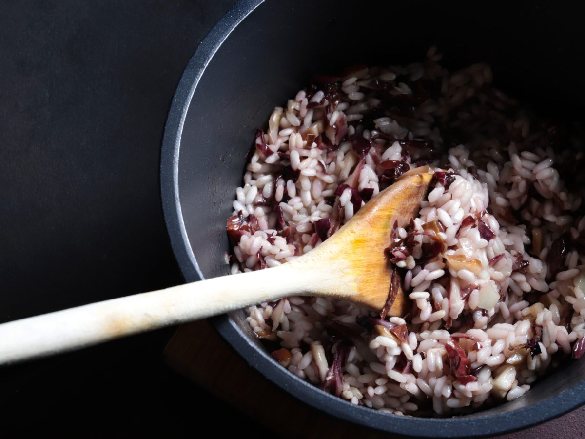 in einem dunklen Topf ist ein italienisches Radicchio-Risotto. In diesem steckt ein großer Kochlöffel au Holz.