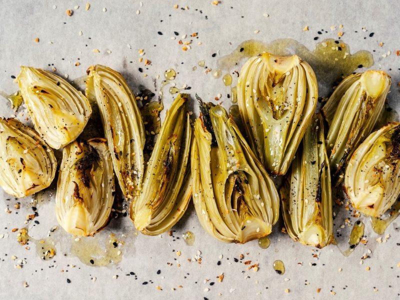 Ein Stück Backpapier, auf dem Stücke von karamellisiertem Fenchel liegen, bestreut mit Pfeffer und Öl.