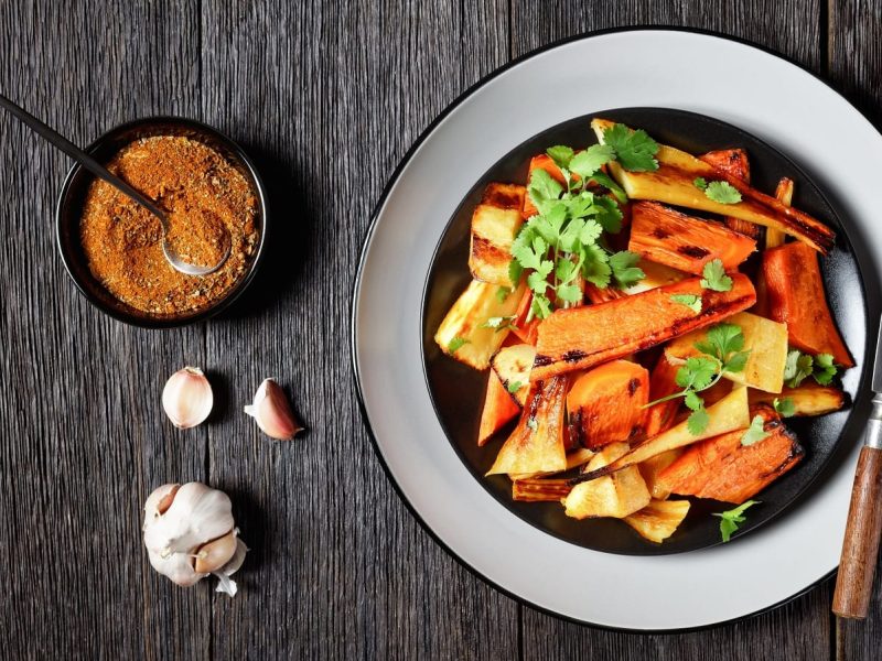 Ein Teller gefüllt mit gebackenen Karotten und Pastinaken, garniert mit frischem Koriander, neben einer Knoblauch-Knolle, einer kleinen Schüssel mit marokkanischen Gewürzen und Messer und Gabel, vor einem Holzhintergrund, von oben.