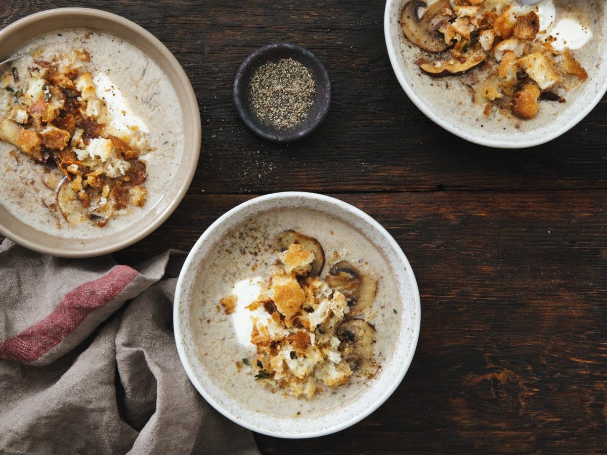 Drei Schüsseln Kartoffelsuppe mit Steinpilzen und Croutons auf dunklem Holz in der Draufsicht.