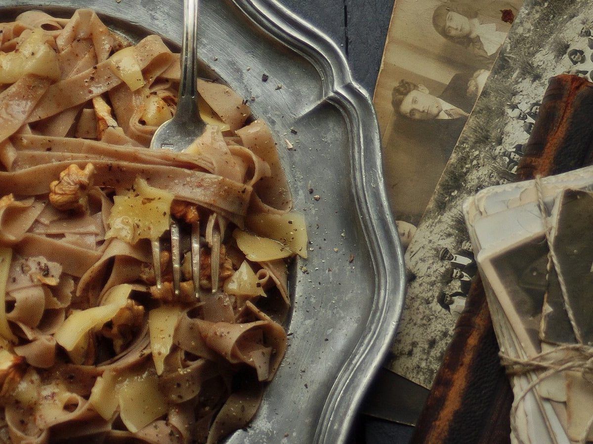 Ein silbener Teller mit einer Portion selbstgemachter Kastanien-Pasta mit Walnusssoße, garniert mit Parmesanscheiben und Walnüssen neben alten Fotografien und Büchern vor einem dunklen Hintergrund, von oben.