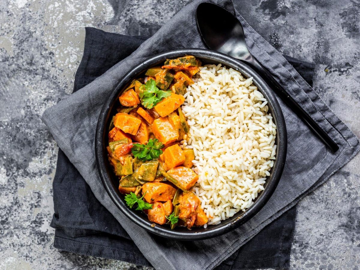 Ein dunkler Teller Reis und Kürbis-Curry mit Lauch steht auf zwei grauen Geschirrtüchern auf einem Marmortisch, Draufsicht.