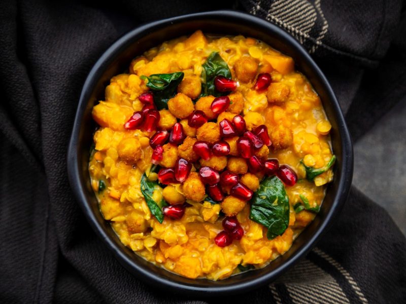 Ein dunkler Teller befüllt mit Kürbis-Linsencurry mit Spinat, Kichererbsen und Granatapfelkernen in der Draufsicht.