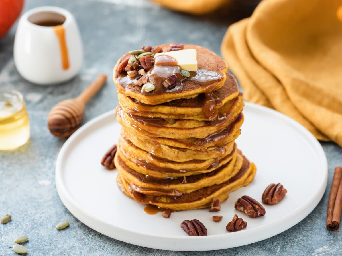 Ein weißer Teller mit gestapelten Kürbis-Pancakes. Darauf Pecannüsse, ein Stück Butter und etwas Sirup. Drumherum sieht man ein Honigglas, Zimtstangen und ein Stück von einem Kürbis.