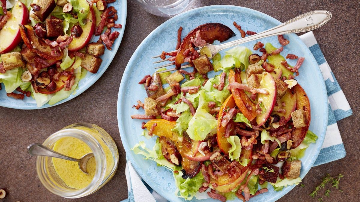 Auf zwei hellblauen Tellern ist ein Kürbis-Salat mit Speck-Croûtons angerichtet. Auf einem Teller liegt eine Gabel. Blick von oben.