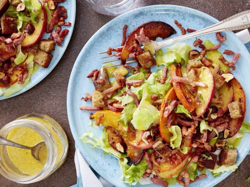 Auf zwei hellblauen Tellern ist ein Kürbis-Salat mit Speck-Croûtons angerichtet. Auf einem Teller liegt eine Gabel. Blick von oben.