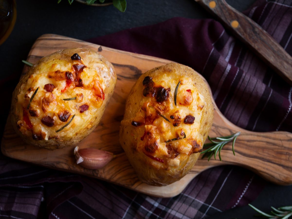 In der Draufsicht sieht man zwei gefüllte Kumpir mit Apfel, Paprika und viel Käse, die auf einem Küchenbrett liegen. Dunkler Untergrund mit einem Geschirrtuch.