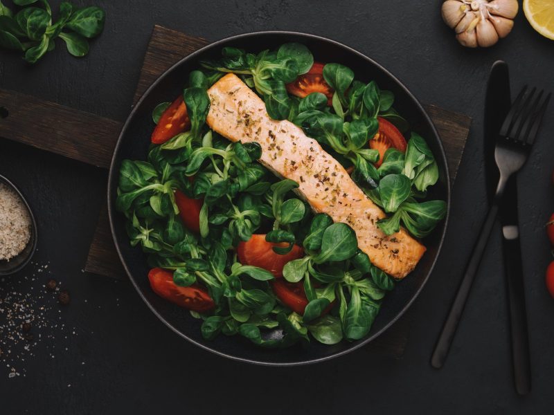 Ein Teller mit Lachs mit Feldsalat und Tomaten auf dunklem Untergrund in der Draufsicht. Drumherum sieht man einzelne Zutaten wie Tomaten, Salz oder eine halbe Zitrone, sowie Besteck.