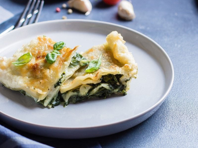 Ein Teller mit zwei Stück Lachs-Spinat-Lasagne bestreut mit frischem Basilikum. Im Hintergrund sieht man unscharf ein paar Knoblauchzehen, Pfefferkörner, Besteck und Geschirrtücher.