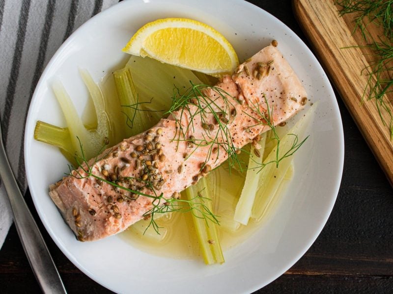 Ein Stück gebackenes Lachsfilet auf Fenchel-Zitronenbett mit Fenchelsamen, Fenchelgrün und frischer Zitrone serviert neben einer Gabel, einem Holzbrett mit Zitronen und Fenchel und einem gestreiften Küchentuch, vor dunklem Hintergrund, von oben.