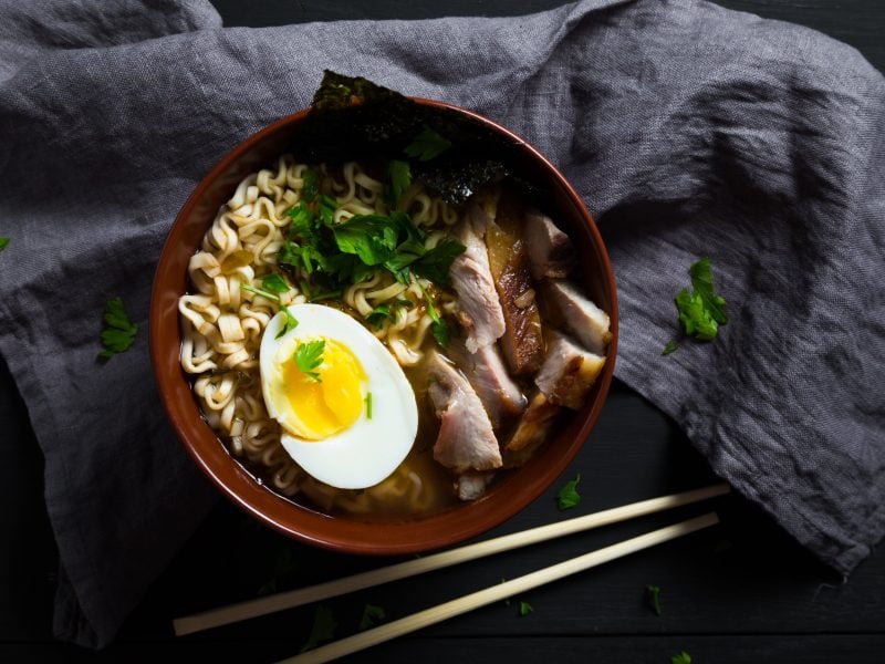 Eine Schüssel mit Miso Ramen mit eingelegtem Schweinebauch, einem halbem Ei, frischem Koriander und einem Noriblatt als Topping, neben einem Paar Essstäbchen, auf einem grauen Küchentuch, vor dunklem Hintergrund, von oben.