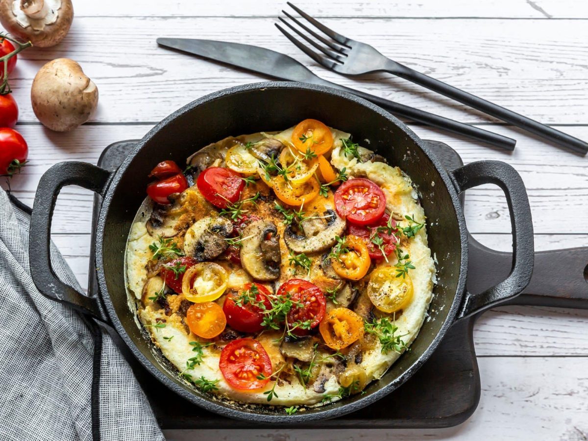 Eine gusseiserne Pfanne mit einem Omelett mit Pilzen und Tomaten sowie Oregano in der Draufsicht auf hellem Untergrund. Daneben liegen Besteck, ein paar Tomaten und Pilze sowie ein Geschirrtuch.