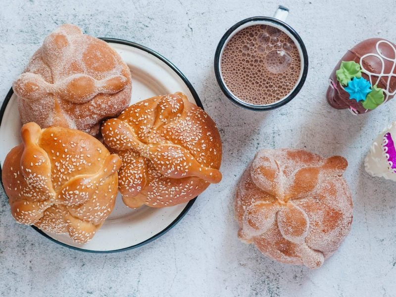 Ein Teller mit drei Pan de Muerto bestreut mit Zucker und Sesam neben einer Tasse heißer Schokolade, einem weiteren Brötchen und zwei Schokoladen Totenköpfen.