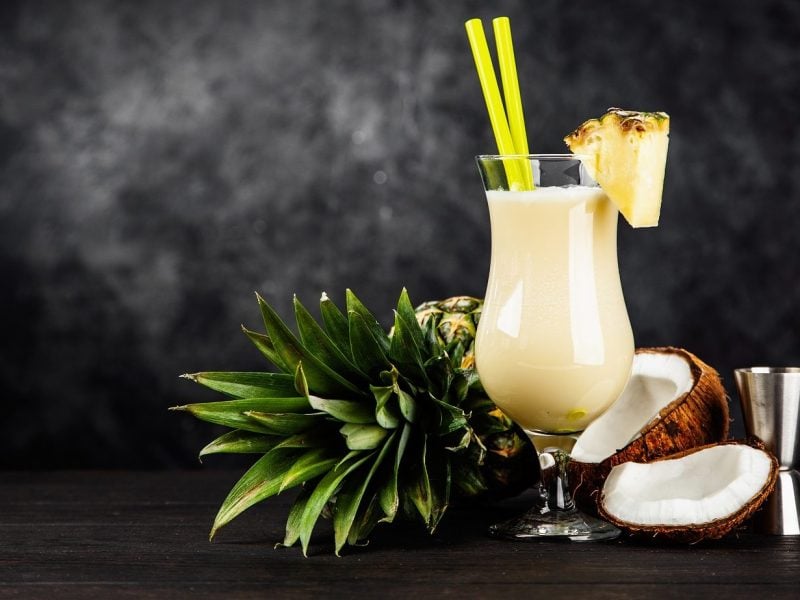 Die Low Carb-Pina Colada stilecht im Glas angerichtet, mit Ananas am Rand verziert und die Kokosnuss als Deko im Hintergrund.