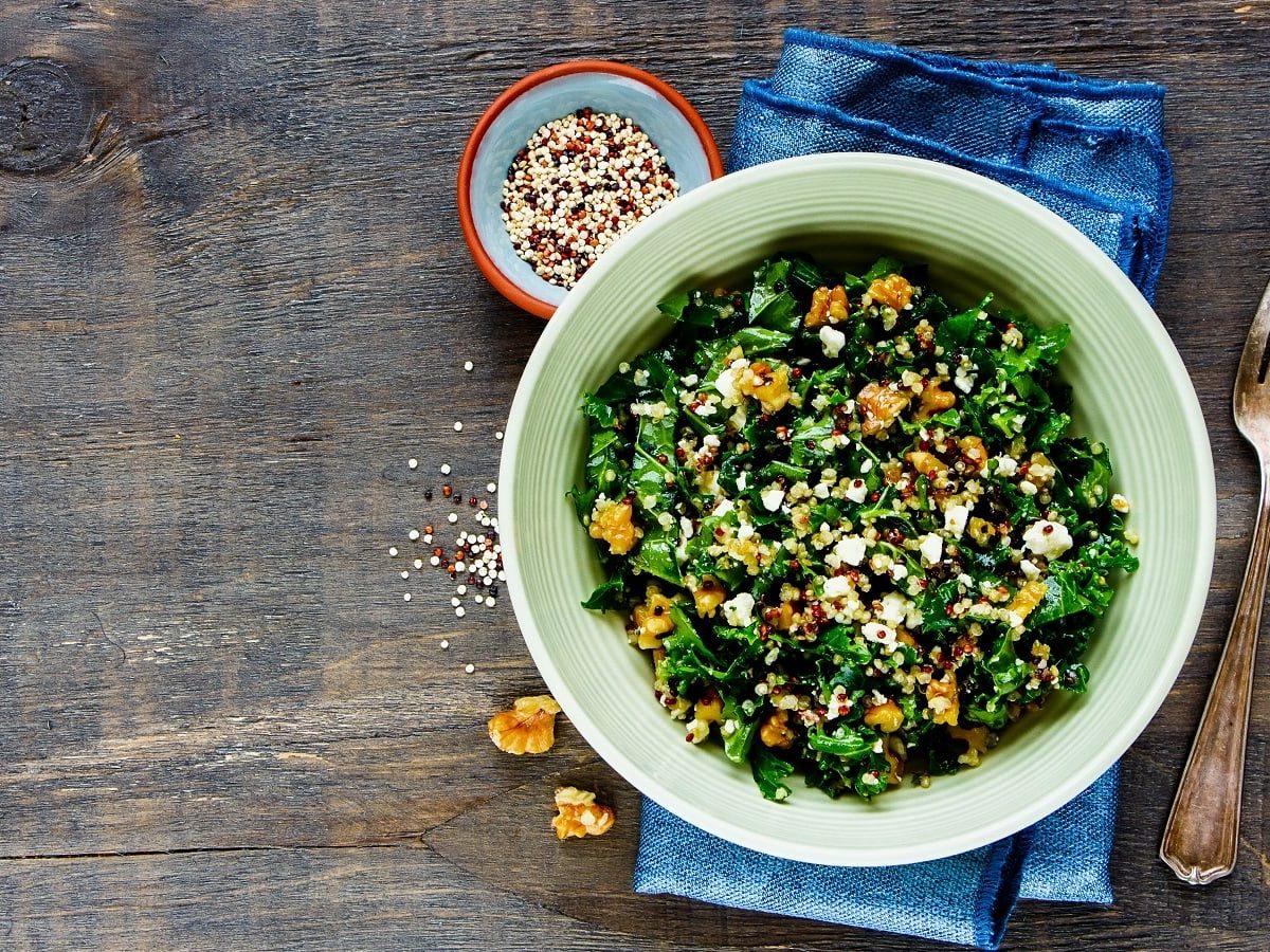 Eine hellgrüne Schüssel gefüllt mit dem Quinoa-Grünkohl-Salat garniert mit gerösteten Walnüssen und zerbröseltem Feta-Käse auf einem blauen Küchentuch neben einer kleinen Schüssel rohen Quinos-Körnern, zwei Walnüssstücken und einer Gabel vor einem Holzhintergrund, von oben.