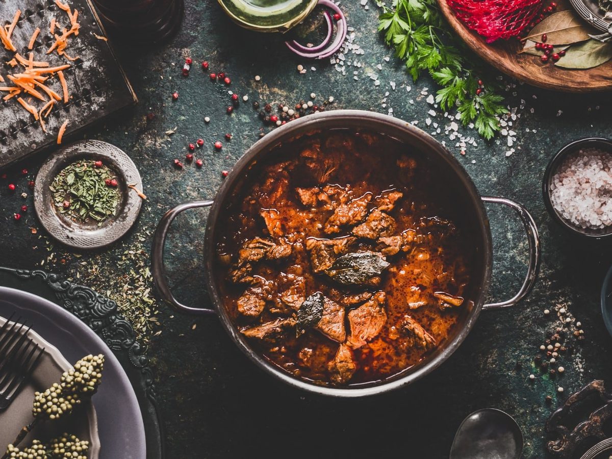 Ein Topf mit Rindergulasch mit Coa in der Draufsicht auf dunklem Untergrund. Drumherum sind eine Vielzahl von Schälchen mit Zutaten zu sehen.